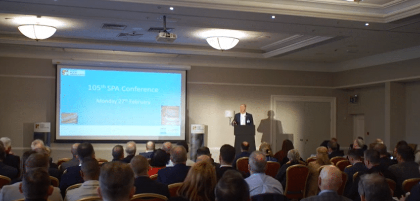 Neil Osment presenting at a conference in front of a large screen and a room full of people
